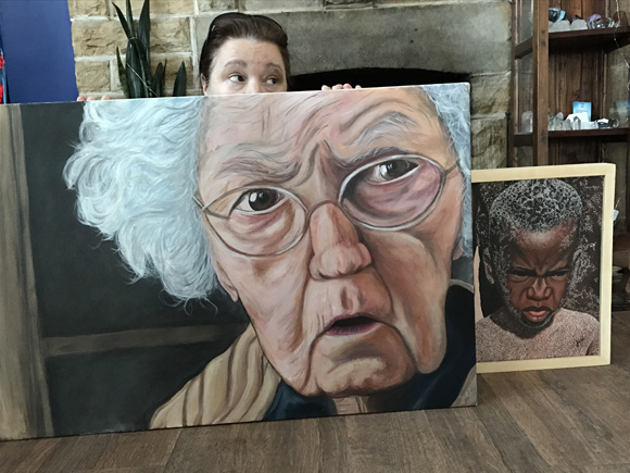 Artist Julie Underwood peaks out from behind two of her portraits currently on display at StoryArt.